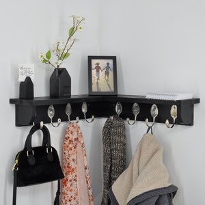 May include: Black corner shelf with hooks and a picture frame. The shelf has a white card with the text "So grateful" and a vase with white flowers. There are also a few items hanging on the hooks, including a black purse, a scarf, and a sweater.