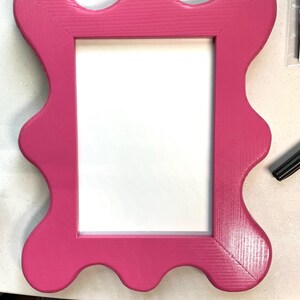 K&ouml;nnte beinhalten: Ein leuchtend rosa Bilderrahmen mit einem wellenf&ouml;rmigen, gewellten Rand. Der Rahmen ist aus Holz und hat einen wei&szlig;en Passepartout.