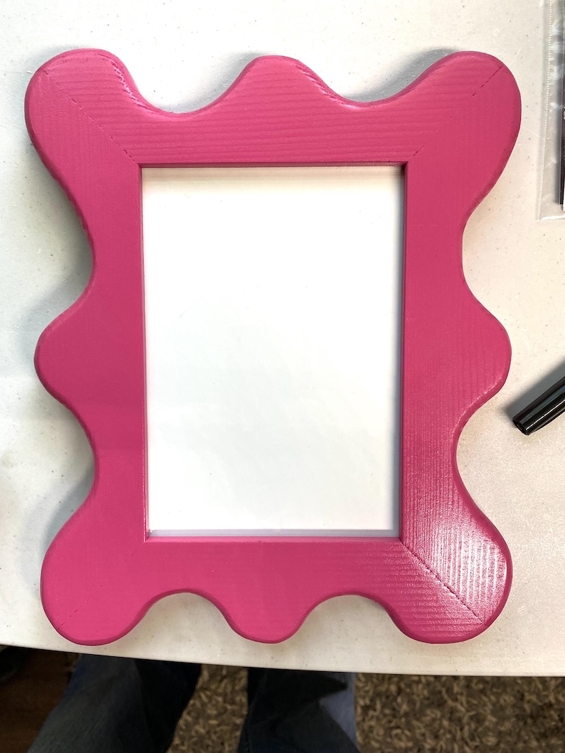 K&ouml;nnte beinhalten: Ein leuchtend rosa Bilderrahmen mit einem wellenf&ouml;rmigen, gewellten Rand. Der Rahmen ist aus Holz und hat einen wei&szlig;en Passepartout.