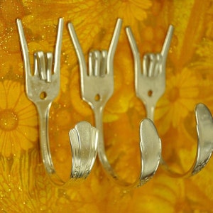 Peut inclure: Trois fourchettes en argent pliées en forme de main faisant le geste rock and roll. Les fourchettes sont montées sur un fond jaune à motif floral.