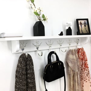 May include: White wall mounted shelf with a white shelf and six silver hooks. The shelf has a black vase with white flowers, a black bird figurine, a black picture frame with a photo of two children, and a small black sign with the text "Love wins, always." hanging on it. There are two scarves and a black purse hanging on the hooks.