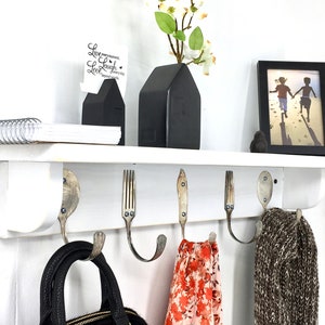 Peut inclure: Étagère blanche avec cinq crochets en fourchettes recyclées. L'étagère est décorée d'un vase noir en forme de maison avec des fleurs blanches, d'une photo encadrée de deux enfants et d'une pile de livres. Le texte "Live every moment, Love every day" est visible sur une carte.