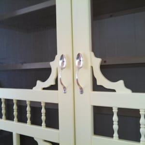 May include: A close-up of a cream-colored cabinet with a screen door. The cabinet doors have decorative cutouts and are adorned with spoon-shaped handles. The cabinet has a traditional design with a balustrade-style lower section.