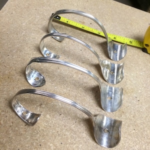 May include: Four silver spoon hooks, bent into a curved shape, with a hole at the top of each hook. The hooks are arranged in a row on a light brown surface.