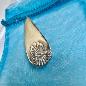 May include: A silver-coloured, decorative, oval-shaped, metal object with a raised, ornate design. The object is sitting on a light blue, sheer fabric.