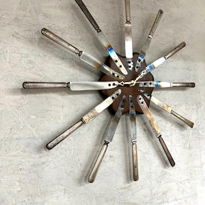 May include: A clock made from repurposed silverware. The clock face is a wooden circle with twelve silver knives arranged in a circle around it. The knives are welded together at the base and the tips point outwards. The clock hands are gold.