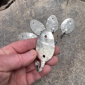 May include: Silverware hooks crafted from repurposed spoons. The hooks have a curved design and are attached to the spoon bowls with a drilled hole. Several hooks are visible, along with screws, suggesting a DIY project or upcycled decor.