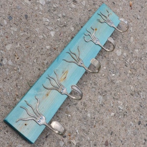 Puede incluir: Un perchero de pared hecho a mano con una base de madera pintada de color turquesa. El perchero presenta cinco ganchos de tenedor plateados, cada uno adornado con un diseño decorativo de pulpo. Los tentáculos del pulpo están hechos con los dientes del tenedor.