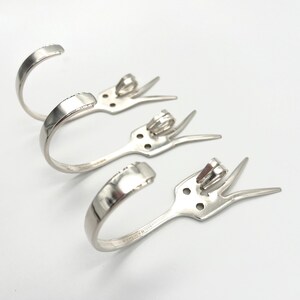 May include: Three silver fork hooks, each with a curved handle and two prongs. The hooks are arranged in a row, with the handles facing the viewer.