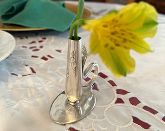 May include: A silver-toned metal bud vase crafted from a repurposed spoon, featuring an upright handle as the vessel and the bowl of the spoon as the base. A single yellow alstroemeria flower with green-tipped petals is placed inside the vase.