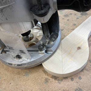 May include: Close-up of a woodworking router in use, shaping a light-colored wooden piece. The router is silver and black, with a clear plastic guard. The wood has a curved shape.