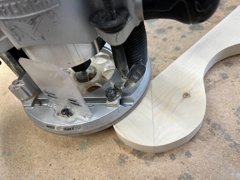 May include: Close-up of a woodworking router in use, shaping a light-colored wooden piece. The router is silver and black, with a clear plastic guard. The wood has a curved shape.
