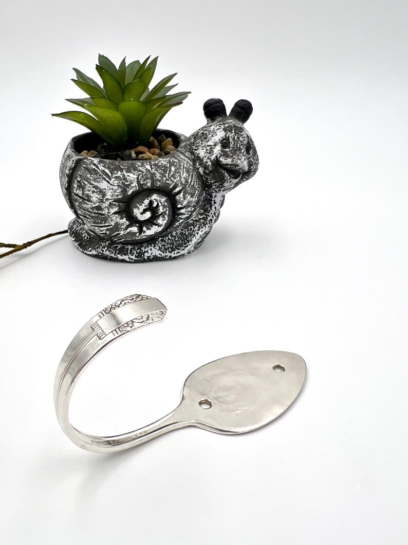 May include: A whimsical snail-shaped planter with a faux succulent and a silver spoon hook. The planter is gray with white accents, and the spoon hook has a curved design with two holes for mounting. The hook is made from a spoon.