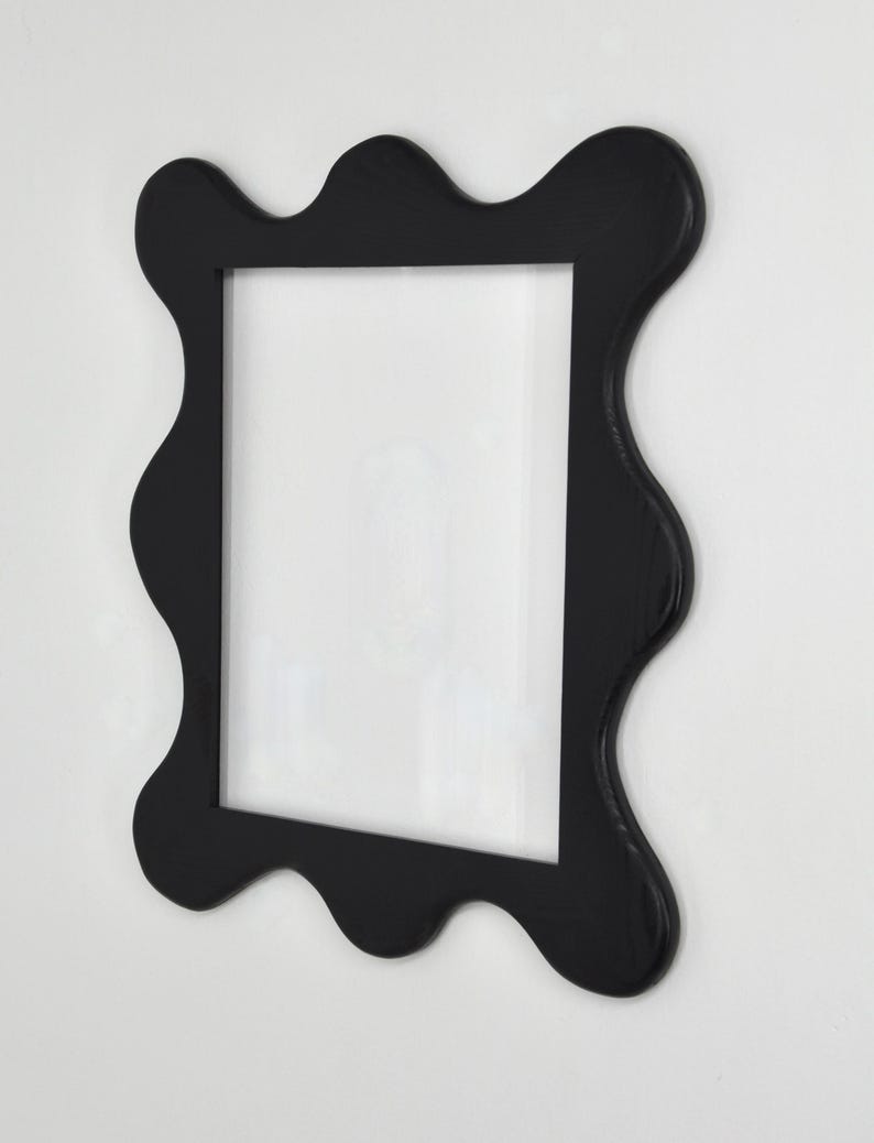 May include: A black picture frame with a wavy, organic edge design. The frame has a rectangular opening and is mounted on a white wall. The frame's unique shape adds a modern touch to the decor.