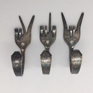 May include: Three silver forks bent into different hand gestures, including peace sign, middle finger, and rock and roll sign. The forks are mounted on a wall as hooks.