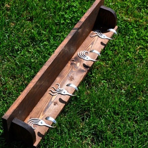 May include: A rustic wooden wall shelf with six decorative metal hooks. The shelf is made of stained wood and has a dark brown finish. The hooks are silver-colored and have a unique, stylized design. The shelf is displayed on a green grassy background.
