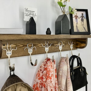 May include: A wooden shelf with five octopus-shaped hooks made from silverware. The hooks are holding a brown jacket, a red and white patterned scarf, a pink and white patterned scarf, and a black purse. The shelf is decorated with a black house-shaped vase, a black bird figurine, and a white card with the text "Live every moment, Laugh every day, Love beyond words."