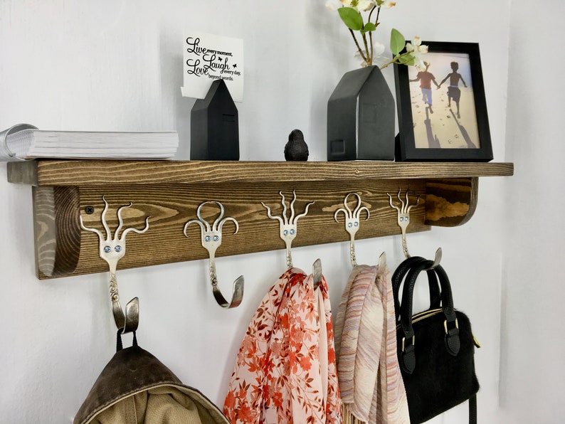 May include: A wooden shelf with five octopus-shaped hooks made from silverware. The hooks are holding a brown jacket, a red and white patterned scarf, a pink and white patterned scarf, and a black purse. The shelf is decorated with a black house-shaped vase, a black bird figurine, and a white card with the text "Live every moment, Laugh every day, Love beyond words."