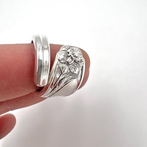 May include: A silver ring with a floral design. The ring features a detailed flower and leaf motif. The band wraps around the finger, creating a unique and elegant look. The ring is made of a shiny, reflective metal.