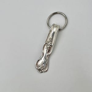 May include: Silverware keychain featuring a spoon with floral detailing. The spoon is attached to a silver metal keyring. The spoon's handle has an ornate design with embossed flowers and scrollwork, creating a vintage aesthetic.