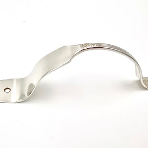 May include: A silver-colored metal handle with a decorative design. The handle is curved and has a hole in the center.
