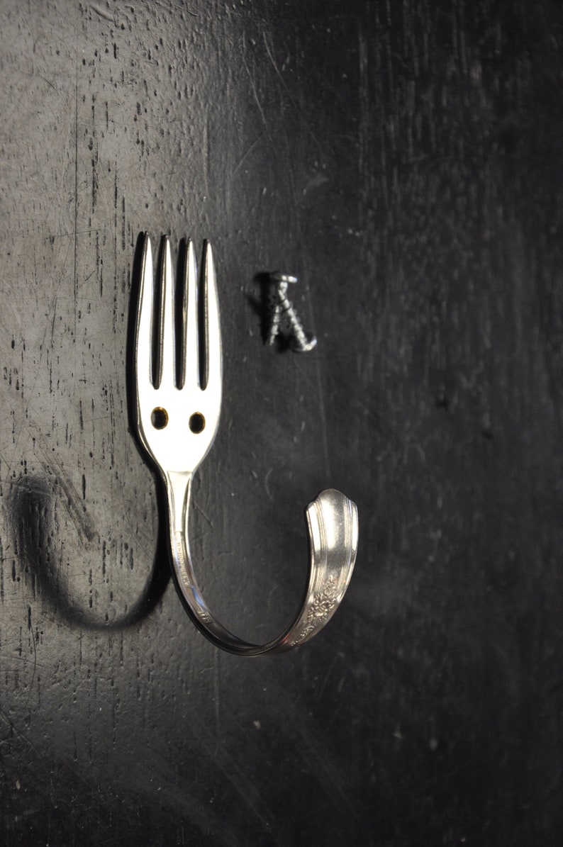 May include: A silver fork bent into a hook shape, mounted on a black surface with two screws.