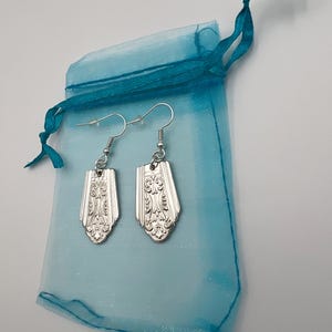 May include: A pair of silver earrings with a decorative design, displayed in a light blue organza bag with a turquoise ribbon.