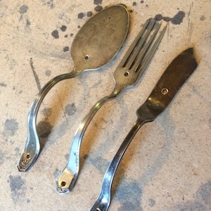 Peut inclure: Trois ustensiles en argent, une cuillère, une fourchette et un couteau, ont été pliés en forme de poignées. Les poignées sont toutes courbées et ont un trou percé en haut.