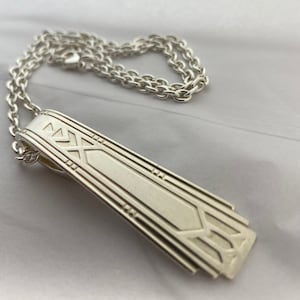 May include: A silver necklace with a long, rectangular pendant featuring an intricate geometric design. The pendant is attached to a silver chain.
