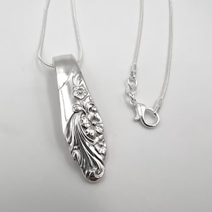 May include: A silver pendant necklace featuring a floral design. The pendant is crafted from a spoon, with intricate details of flowers and leaves. The necklace has a delicate chain with a lobster clasp.