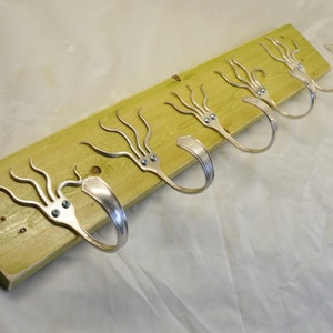 Puede incluir: Un perchero de pared hecho a mano. El perchero está hecho de una tabla de madera verde claro con cinco ganchos de tenedor plateados. Los tenedores están sujetos a la madera con tornillos pequeños y tienen un diseño artístico único. Ideal para colgar abrigos, sombreros u otros artículos.