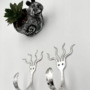 May include: Two silver metal wall hooks with a fork-like design, featuring a face and tentacle-like extensions. A snail-shaped planter with a succulent plant sits above the hooks. The planter is gray and white.