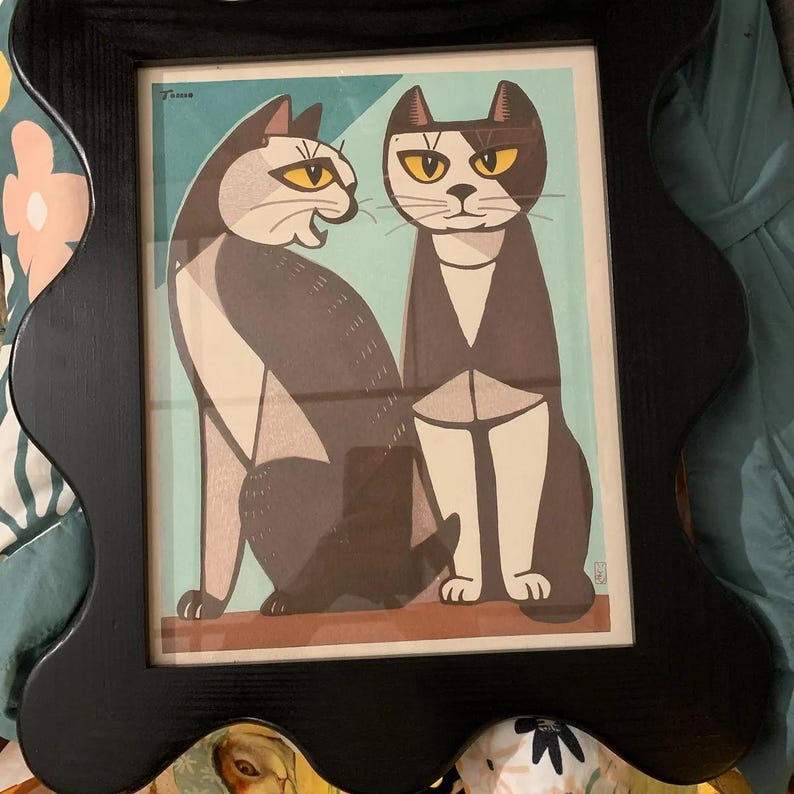 May include: A framed art print featuring two stylized cats with yellow eyes. One cat is gray and white, the other is black and white. The print is in a black wavy-edged frame. The background is a light blue.
