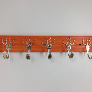 Puede incluir: Un perchero de pared hecho a mano con cinco ganchos. El perchero está hecho de una tabla de madera naranja teñida con ganchos de tenedor plateados. Los tenedores están unidos a la tabla con tornillos y están espaciados uniformemente.