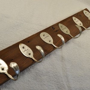 Peut inclure: Un porte-manteau en bois avec six crochets fabriqués à partir de cuillères réutilisées. Chaque cuillère porte un nom gravé : "Ben", "Nadia", "Simone", "Lila", "Finn" et "Liam".