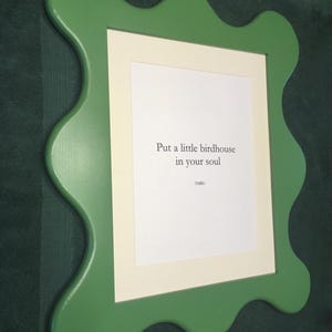 May include: A green, wavy-edged picture frame with a white mat and a printed message: "Put a little birdhouse in your soul." The frame is mounted on a dark green wall. The text is in a simple, sans-serif font.