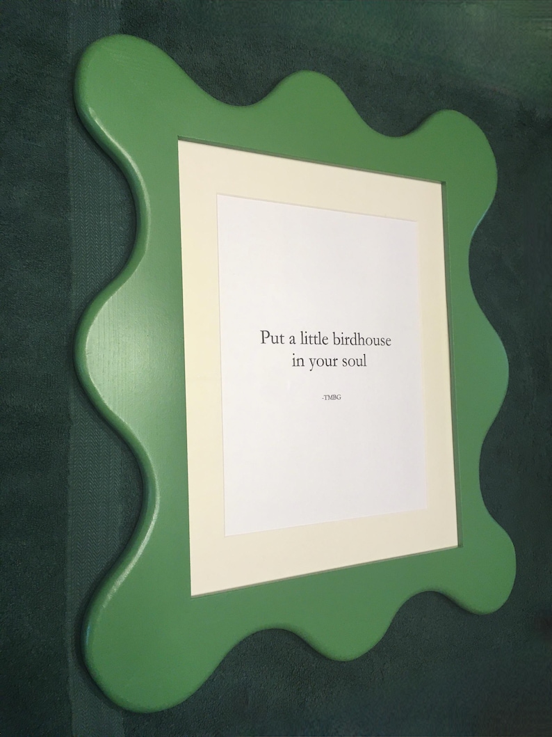 May include: A green, wavy-edged picture frame with a white mat and a printed message: "Put a little birdhouse in your soul." The frame is mounted on a dark green wall. The text is in a simple, sans-serif font.
