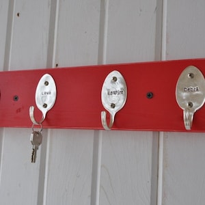 May include: A red wooden key holder with four metal spoon hooks. Each spoon has a word stamped on it: "Create", "Love", "Inspire", and "Debra". A set of keys hangs from the second hook. The key holder is mounted on a white paneled wall.