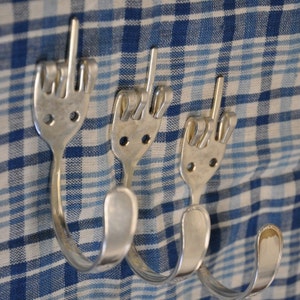 May include: Three silver metal coat hooks shaped like middle fingers, mounted on a blue and white checkered fabric background.