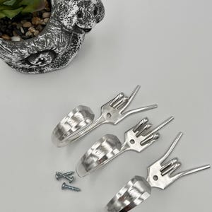 May include: Three silver-colored fork-shaped napkin rings with a rock-and-roll hand gesture design. The napkin rings are next to three small screws. A snail-shaped planter with a succulent plant is in the background.