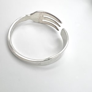 May include: A silver-toned metal ring crafted from a bent fork. The circular band features the tines of the fork at one end and the handle at the other, creating an open-ended design. The metal surface has a smooth, polished finish.