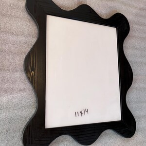 May include: Black wooden picture frame with a unique wavy edge design. The frame has a white interior and the handwritten text "11x14". The frame is designed to hold an 11x14 inch photo or artwork.