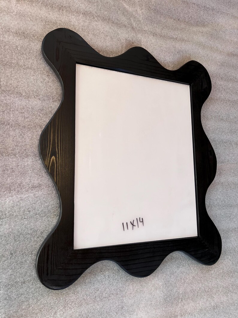May include: Black wooden picture frame with a unique wavy edge design. The frame has a white interior and the handwritten text "11x14". The frame is designed to hold an 11x14 inch photo or artwork.