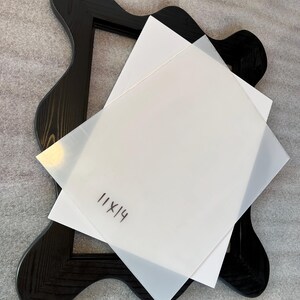 May include: A black wooden frame with a unique, wavy design. Two sheets of white paper, one translucent, are layered on top. The text "11x14" is handwritten on the top sheet. The frame is set against a neutral gray background.