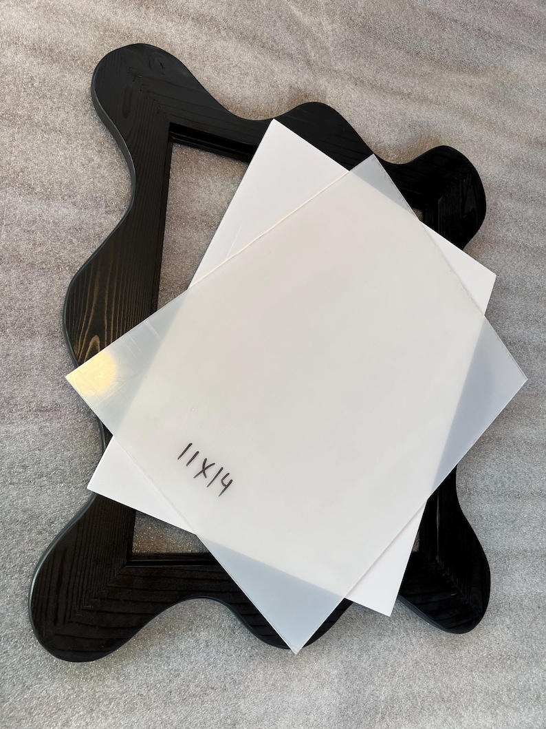 May include: A black wooden frame with a unique, wavy design. Two sheets of white paper, one translucent, are layered on top. The text "11x14" is handwritten on the top sheet. The frame is set against a neutral gray background.