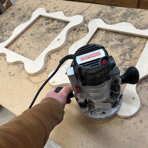 May include: A Craftsman router is used to shape the edges of two unfinished wooden picture frames. The frames have an ornate, wavy design. The router is black and silver with a red power button. The frames are light-colored wood.