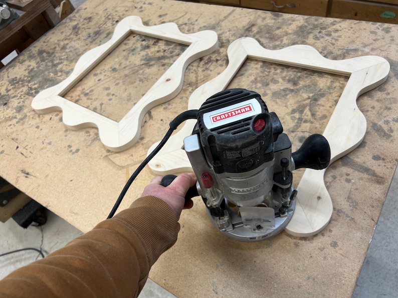 May include: A Craftsman router is used to shape the edges of two unfinished wooden picture frames. The frames have an ornate, wavy design. The router is black and silver with a red power button. The frames are light-colored wood.