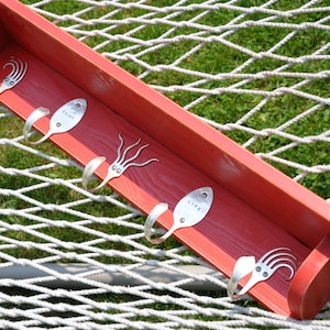May include: A red wooden shelf with six metal hooks made from repurposed silverware. The hooks are attached to the shelf, which is resting on a white rope net. Some hooks are stamped with the words "ENJOY" and "LIFE".