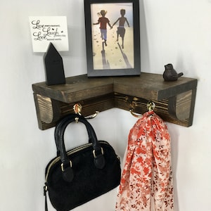 May include: A corner shelf unit made of dark stained wood, featuring two shelves and two brass hooks. A black handbag and a floral scarf hang from the hooks. A framed photo, a small black house, and a bird figurine are on the shelves. The text "Live every moment, Laugh every day, Love beyond words" is on a white card.