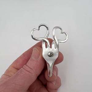 May include: A silver fork with two heart-shaped designs at the top. The fork's tines are connected to a handle, and the hearts are formed by curving the metal. The fork is held against a white background.